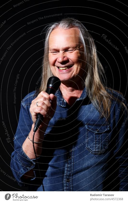 elderly man with long gray hair sings into a microphone long hair song stage black background music old senior male singer happy leisure fun mature concert