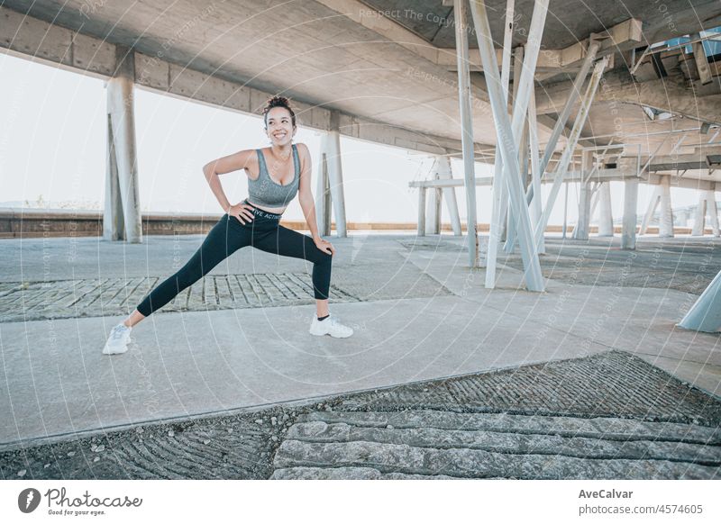 Smiling young african woman practicing yoga, stretching legs over the mat, health instructor. Working out, wearing sportswear, black pants and top, urban outdoor city full length,looking at the camera