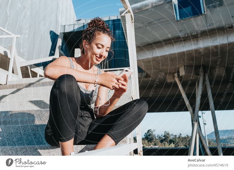 Pretty african american young sportswoman with sport clothes and elastic band looks at smart phone resting at training near modern building. Sportswear, smart phone, headphones.Active lifestyle