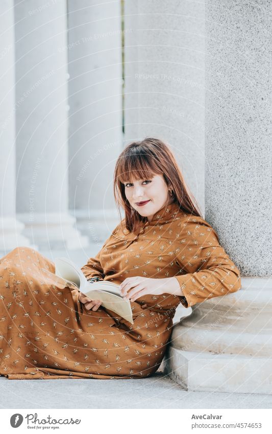 Young woman reading book and smiling to camera while sitting in the floor in an architectonic place. Happy while learning and reading adventures. Copy space. 80s vibes. Trendy styling dress