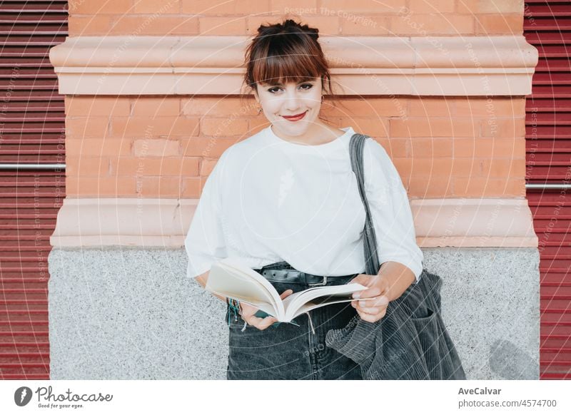 Young happy woman sitting outside the train station, brick wall, reading a book and smiling to camera happy. Waiting for his train transport to arrive, white tshirt with copy space for text