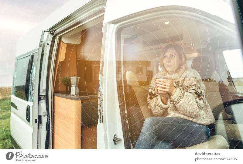 Woman with a cup sitting in the front seat of a camper van woman motorhome cold warming hands coffee sunlight dawn copy space thoughtful winter morning
