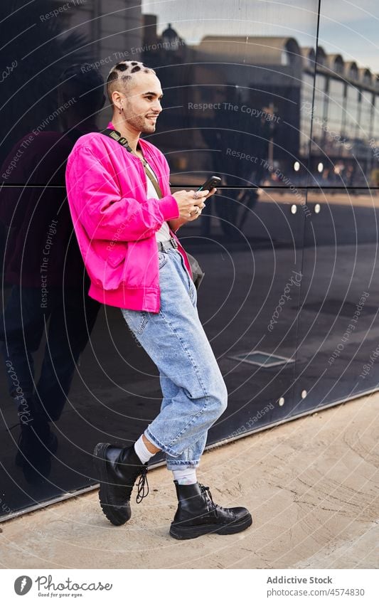 Stylish young man browsing smartphone near reflecting wall eccentric style street using cool message modern reflection male unusual cellphone dyed hair surfing