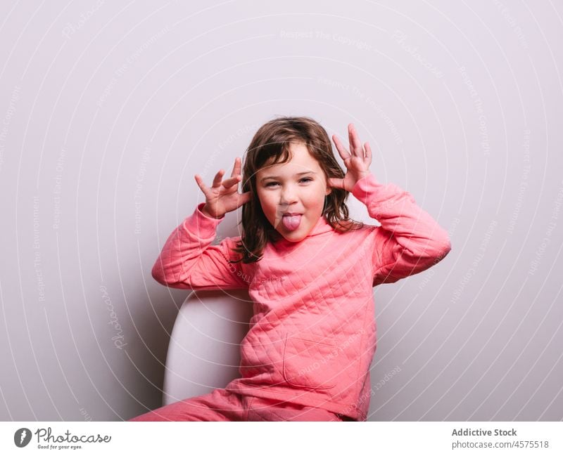 Funny little girl making face grimace tease show tongue funny gesture happy chair sit kid child portrait cheerful tongue out wall playful joy childish make face