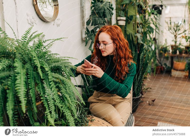 Positive red haired female using a smartphone woman botanic grow plant garden positive foliage cellphone botany potted self portrait charming eyeglasses redhead