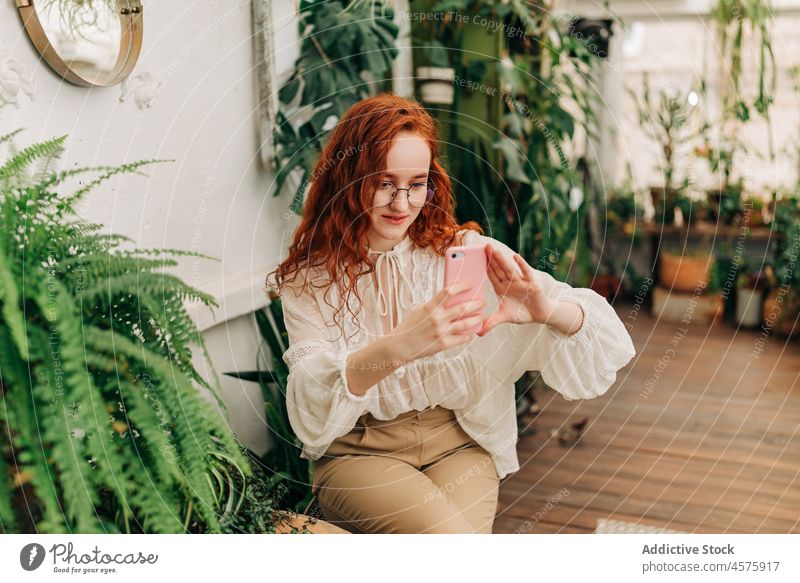 Positive red haired female taking selfie on smartphone woman using botanic grow plant garden positive foliage cellphone botany potted self portrait charming
