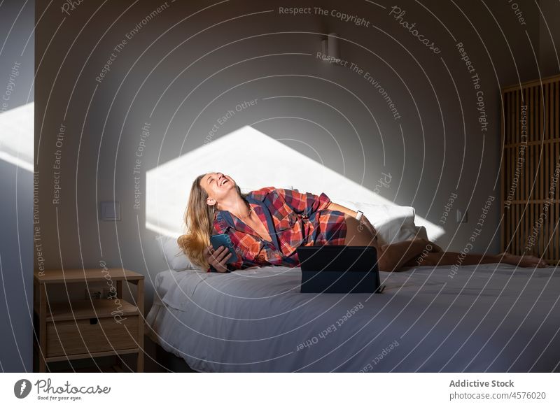 Cheerful woman lying on bed with smartphone and tablet laugh happy using cellphone chill bedroom rest browsing comfort gadget cheerful relax optimist positive