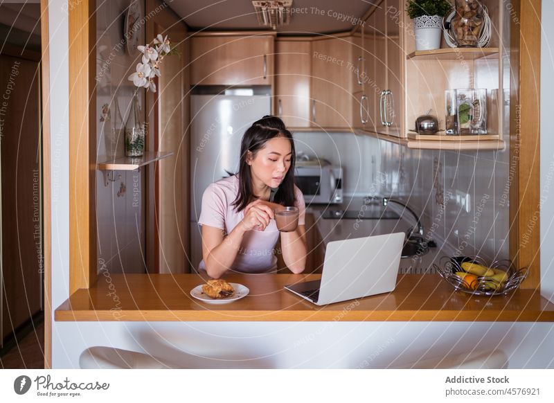 Asian woman with beverage browsing laptop counter kitchen online breakfast hot drink internet pastry food bakery morning female delicious tasty appetizing