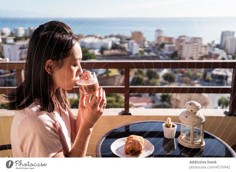 Cheerful Asian woman with beverage on balcony drink breakfast terrace morning hot drink cityscape refreshment female building fence table delicious tasty