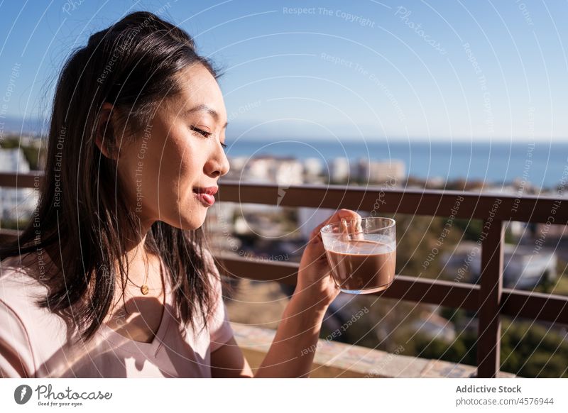 Cheerful Asian woman with beverage on balcony drink breakfast terrace morning hot drink cityscape female building fence delicious tasty appetizing railing glass