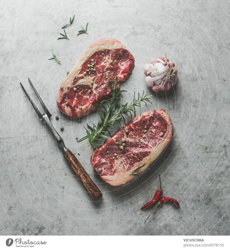 Raw marble rib eye steak with garlic, chili and meat fork on grey concrete kitchen table. Cooking preparation with meat and flavorful seasoning. Top view. raw