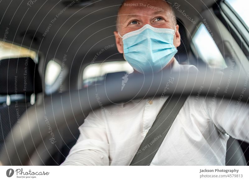 A man in a protective mask driving a car, steering wheel in the foreground driver taxi uber ride surgical mask viral adult belt lifestyle delivery inside