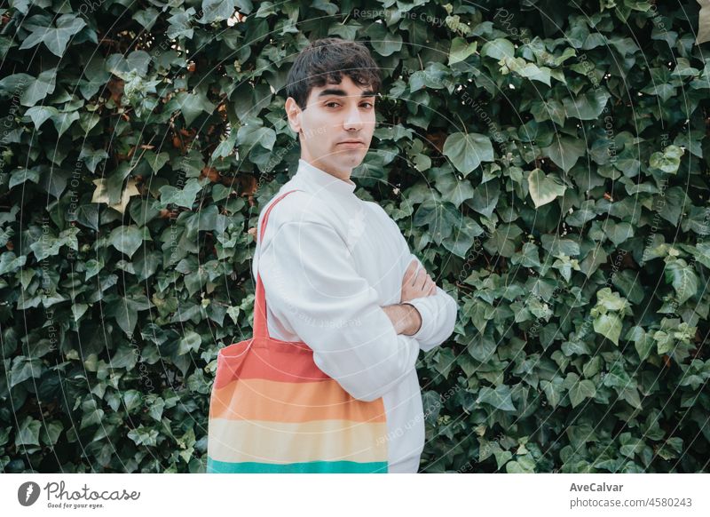 Serious young spanish man looking at camera with a lgtbi tote bag looking at camera smiling on the street, wearing white sweater. Gay pride movement LGBTI, and modern style. Gay concept