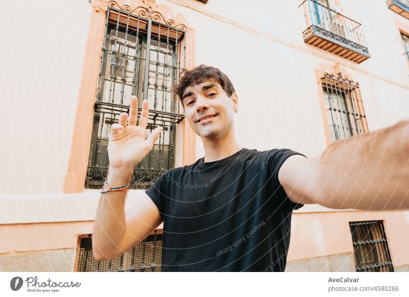 Happy young man wearing shirt taking a selfie in the city during a sunny day and smiling. Copy space, Good vibes concept. Video call saluting. Travel selfie in the europe streets