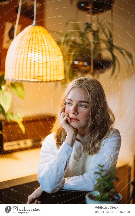 Young girl sitting at desk under illuminated chandelier woman lamp room light home blouse hang flower design plant glow potted shiny modern furniture interior