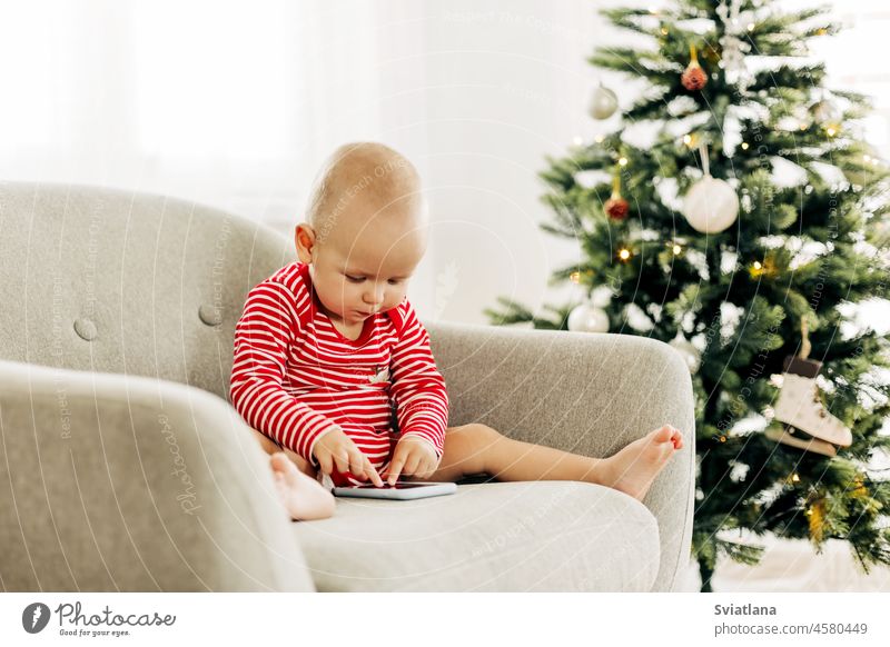 Happy boy kid watching cartoons on a smartphone in a chair next to a Christmas tree happy christmas baby new year playing christmas tree sitting home cute child