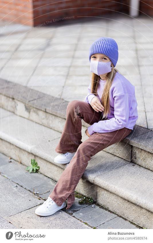 Cute little caucasian girl eight years old with blonde hair and protective face mask walking outdoor. Kid wearing stylish shirt and knitted hat violet color. Trendy color of the 2022 year very peri