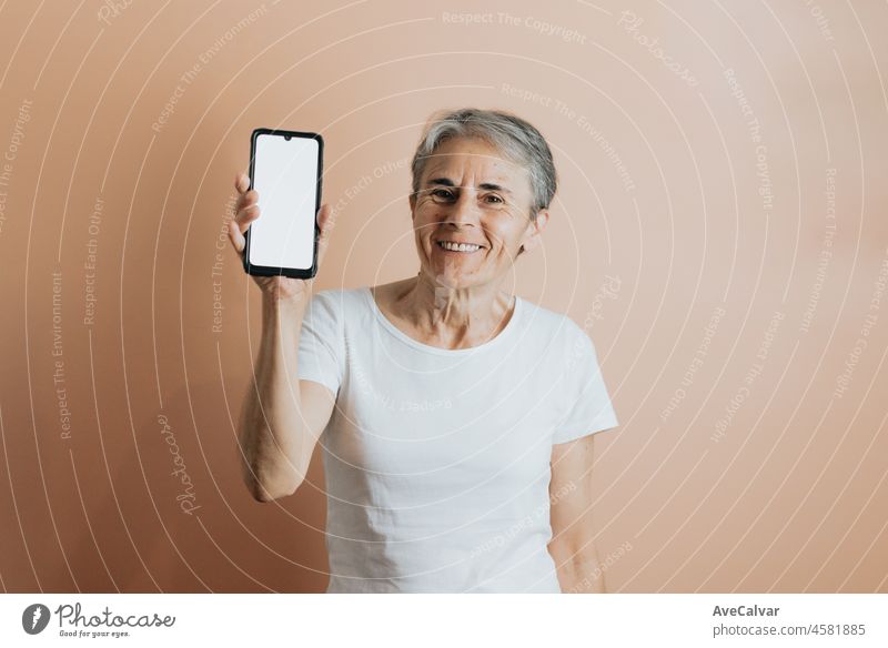 Old senior woman showing to camera a mobile phone smart phone with copy space to put text ad. Senior people colourful background. White tshirt copy space