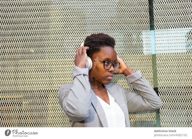 Young pensive African American woman listening to music in headphones grid song fence wireless thoughtful gadget device street positive sound melody meloman