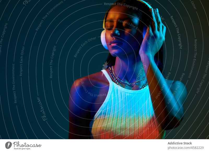 Black Brazilian woman listening to music under fluorescent light style trendy studio headphones song meloman ultraviolet black ethnic eyes closed african modern