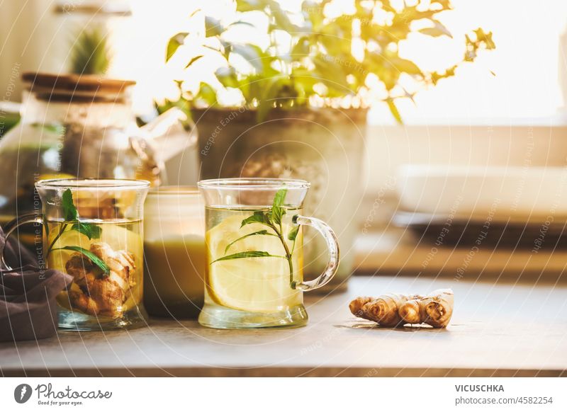 Two glass cups with fresh lemon and ginger tea on kitchen table with ginger root, herbs and kitchen utensils at window background. Healthy homemade tea with vitamin c in wintertime. Front view.