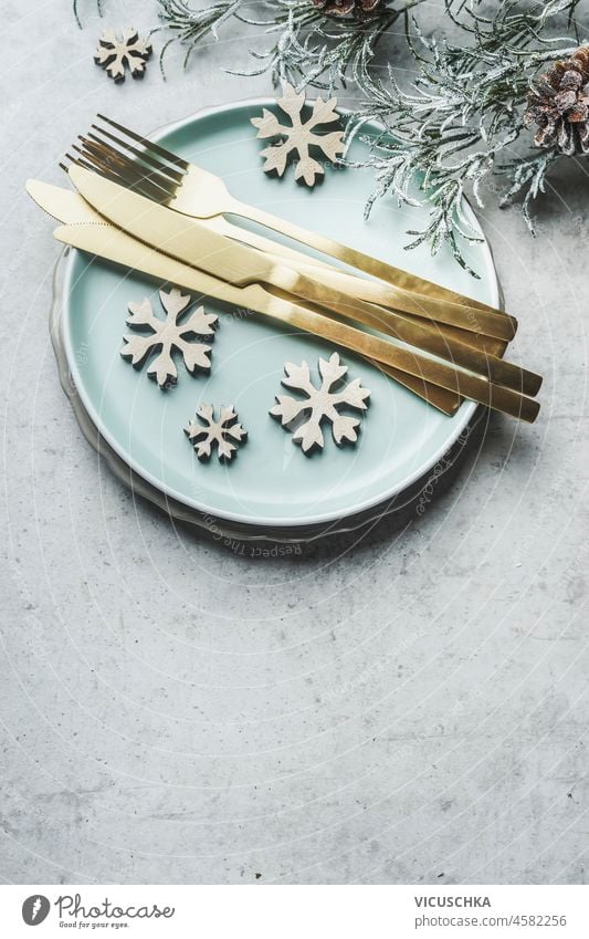 Christmas table setting with pale blue plate, golden cutlery, wooden snowflakes, fir green and pine cone on grey concrete table. Festive table decoration for Christmas dinner. Top view with copy space