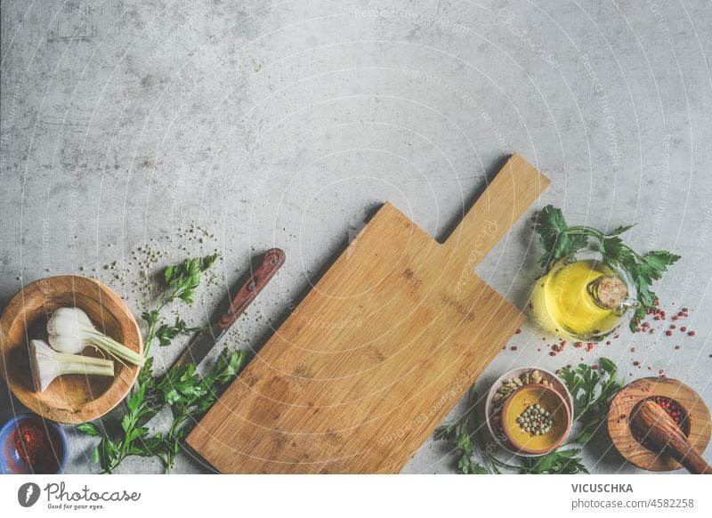 Food background with wooden cutting board, fresh herbs, garlic, olive oil, mortar and pestle,  kitchen knife and spices on grey kitchen table. Cooking preparation with flavorful ingredients, Top view.