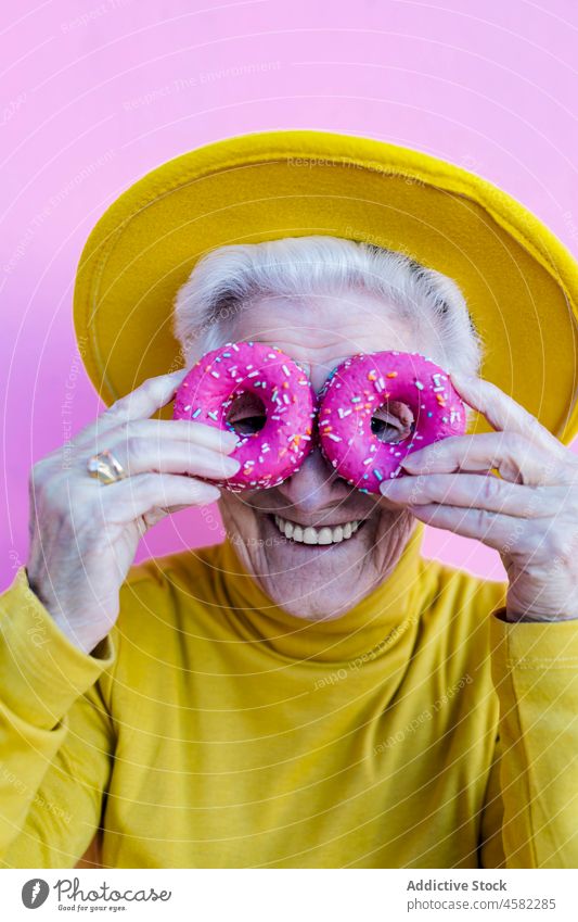 Aged woman covering eyes with doughnuts cover eyes dessert having fun carefree elderly old retire portrait sweet wall style aged happy female hat smile