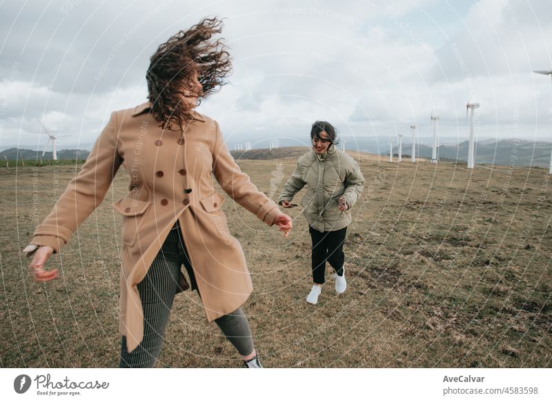 Two young women, on winter cloths during a windy day, enjoying a day while having fun running togueter between electric windmill . Lifestyle. Hipster and modern style. Cinema style.