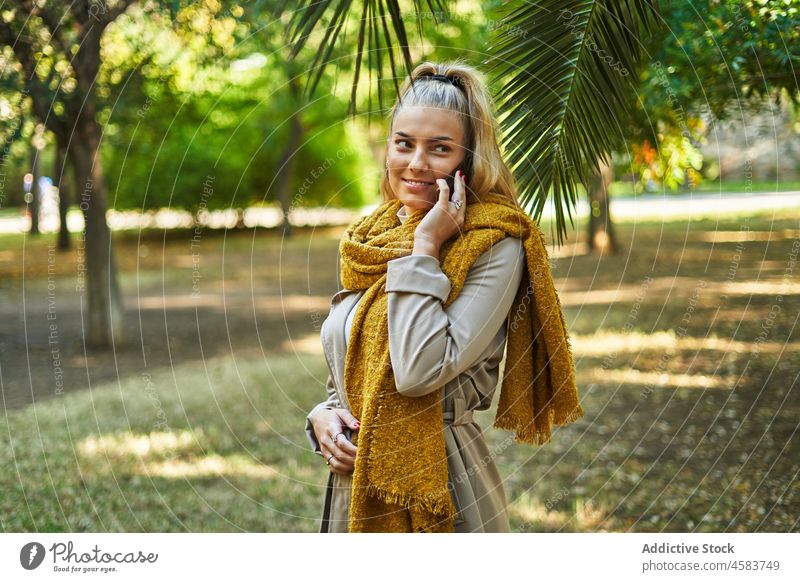 Smiling female in warm scarf talking on cellphone in park woman smartphone speak mobile coat conversation positive outerwear blond using stroll communicate palm