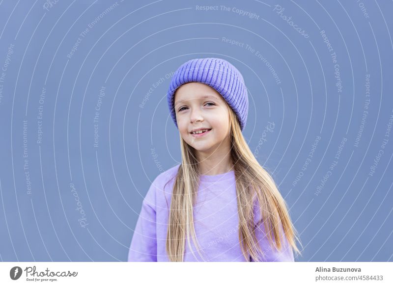Cute little caucasian girl eight years old with blonde hair smiling outdoor. Kid wearing stylish shirt and knitted hat violet color. Trendy color of the 2022 year very peri