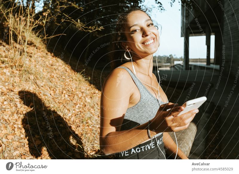 Smiling fit african arab woman in tank top and leggins uses his phone while resting sitting outdoors during a sunset,outdoor stadium going to practice yoga to relax after stressful day,with copy space