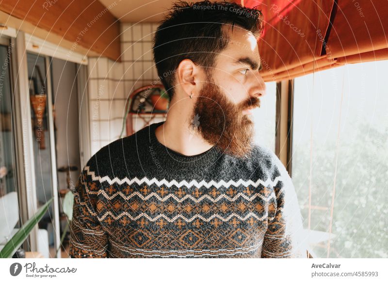 Depressed young bearded handsome man near window at home feeling sad, tired and worried, suffering depression in mental health, problems and broken heart. Psychology problems among men. Colour image