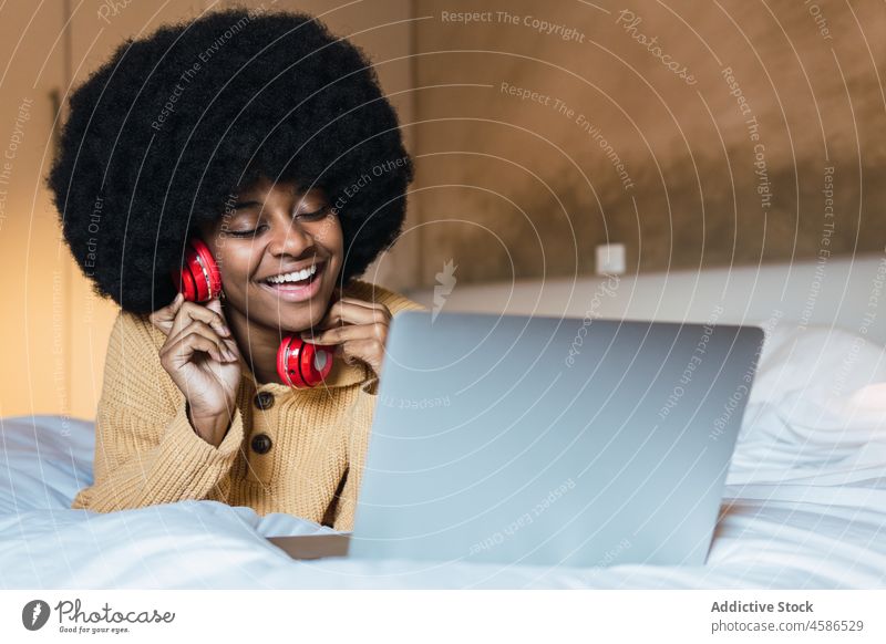 Smiling black woman listening to music with laptop in bed earbuds using home relax rest weekend female african american afro device wireless comfort young