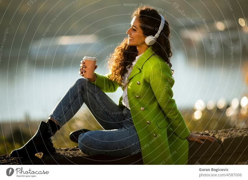 Pretty young woman listening music with smartphone and takeaway coffee by the river on a sunny autumn day pretty attractive caucasian outdoor beautiful