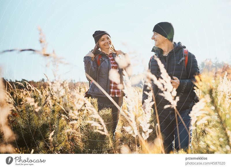 Couple having fun while vacation trip. Hikers with backpacks on way to mountains. People exploring nature walking through tall grass along path in meadow on sunny day