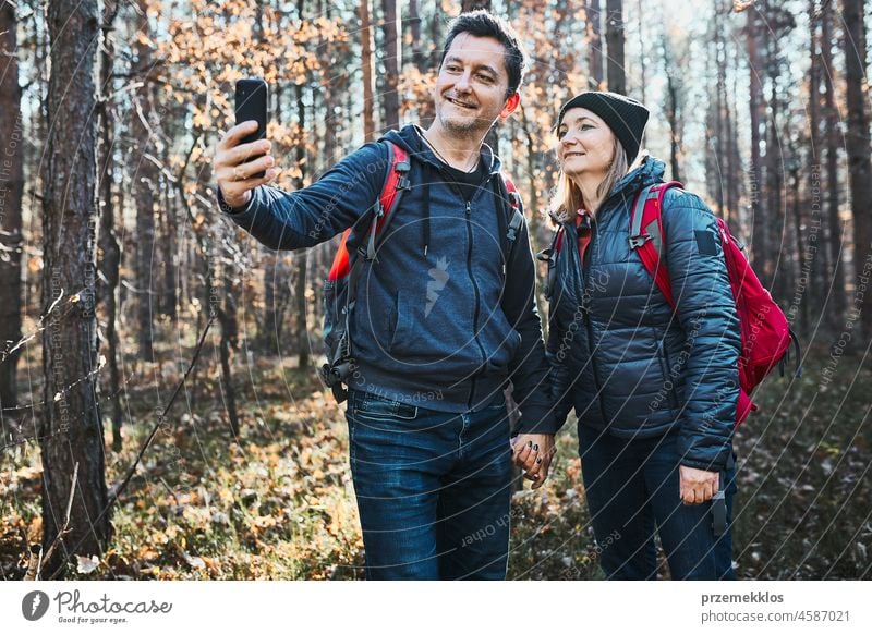 Couple taking selfie photo while vacation trip. Hikers with backpacks walking on path in forest on sunny day hiking adventure travel greeting waving gesture