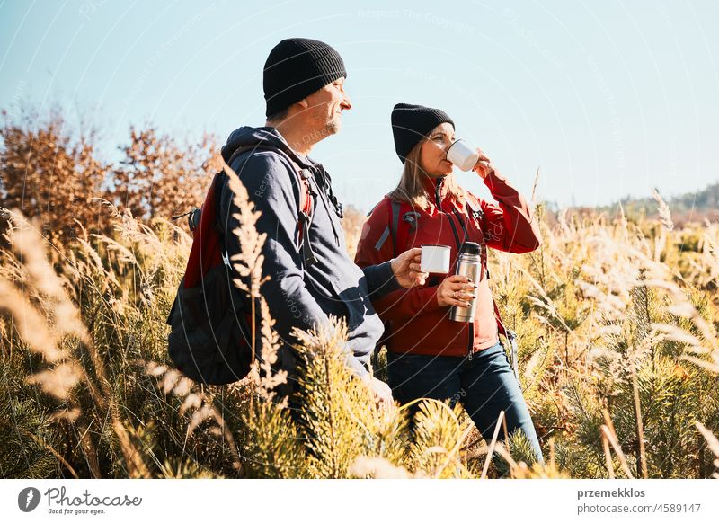 Couple relaxing and enjoying the coffee during vacation trip. People standing on trail drinking coffee. Friends with backpacks hiking through tall grass along path in mountains
