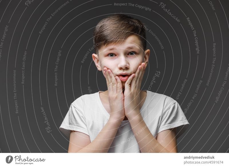 surprised boy holding his head on face isolated on gray background with copy space for text, child in shock, toothache. teenager in white t shirt male emotion