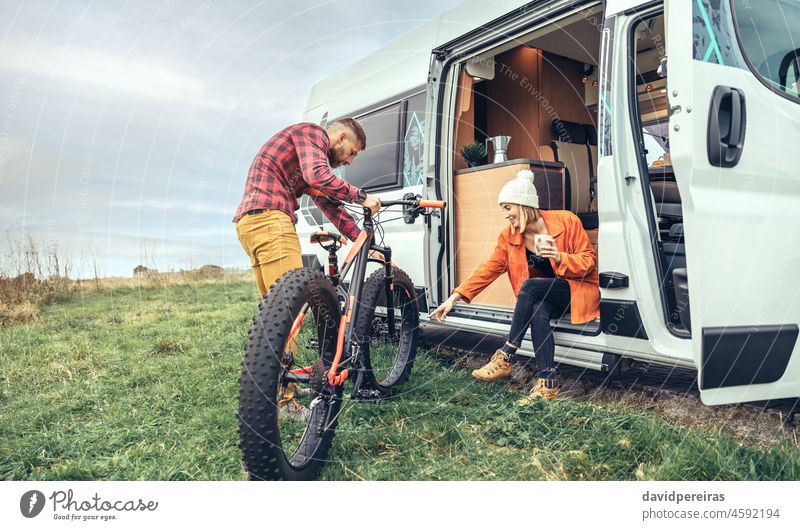 Woman drinking coffee sitting at the door of a campervan and man checking fat bike woman motorhome cold winter copy space smiling preparing morning looking