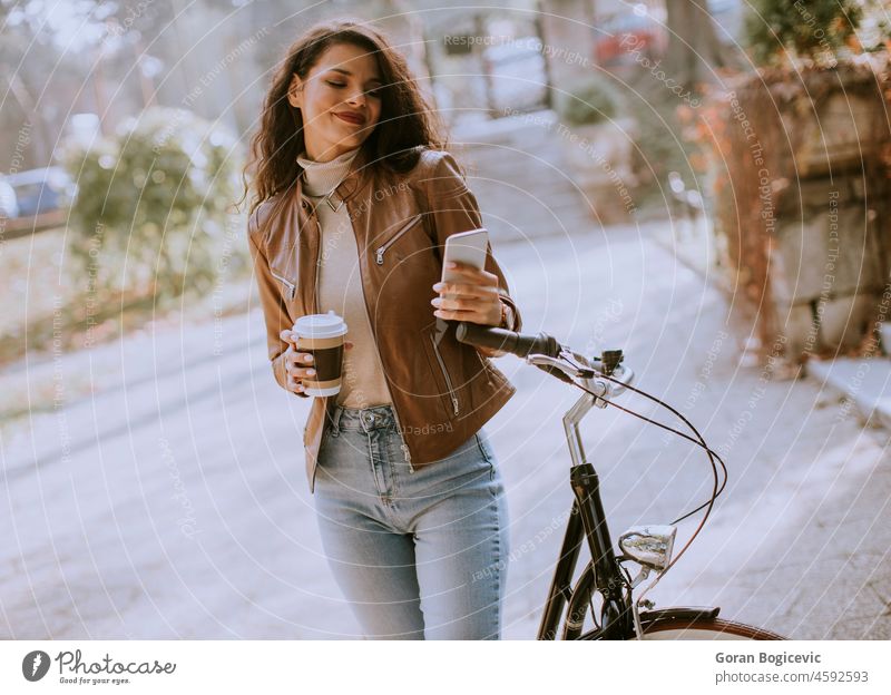 Young woman with mobile phone drink coffee to go by the bicycle on autumn day adult attractive beautiful bike biker business casual caucasian cellphone city