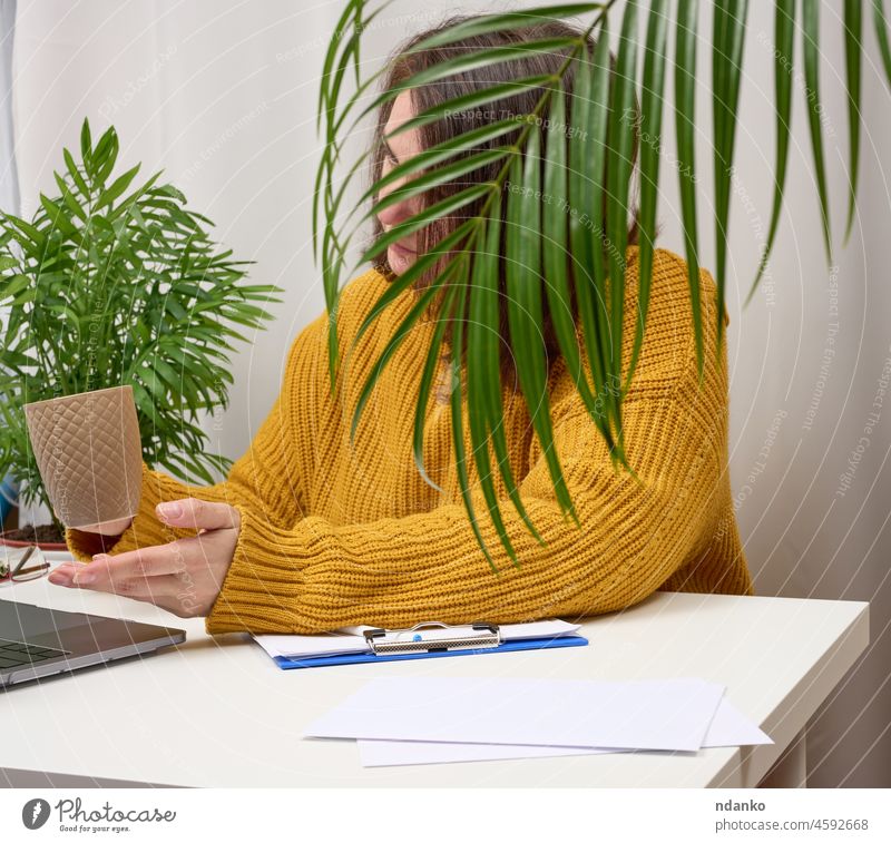 woman 40 years old in an orange sweater sits at the table and holds a cup of coffee in her hand. She uses a laptop, telecommuting, freelancing 40-49 years adult