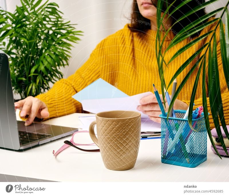a woman in an orange sweater sits at a white table and works at a laptop. Freelancer at remote work, salesperson's home office online adult business casual
