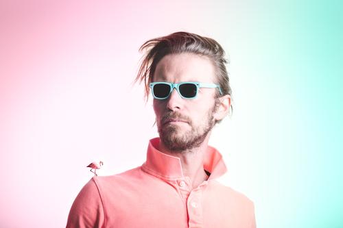Portrait of bearded man in pink polo shirt with light blue sunglasses having a flamingo on his shoulder on pastel background adult color colour long hair look