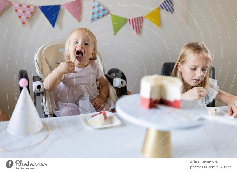 Birthday party at home. Happy little caucasian girl and cute baby celebrating first birthday at home. Candid lifestyle portrait of Kid blowing candles on cake. Living room decorated with flags.