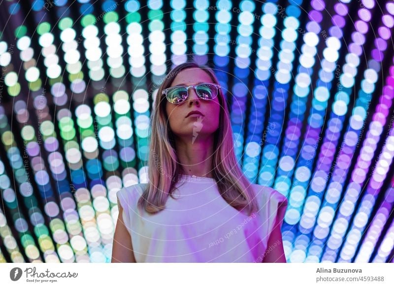 Teen hipster girl in stylish glasses standing on blue tunnel with neon light wall background, female teenager fashion model pretty young woman looking at night club city light glow
