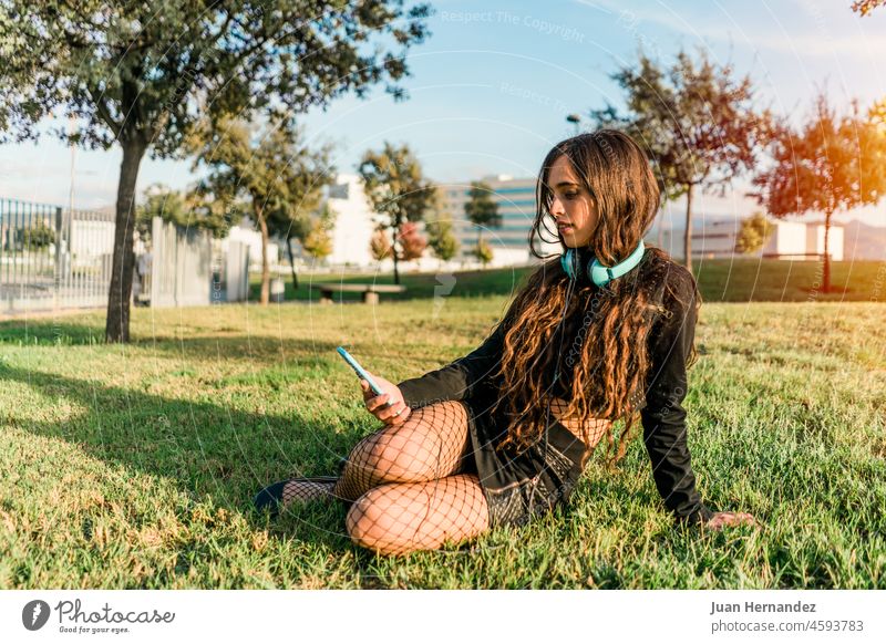 young girl sitting on the grass in a park looking at the smartphone with headphones pretty smart phone earphones mobile phone telephone carefree relaxed