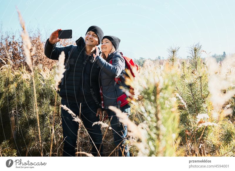 Couple having video call sending greetings from vacation trip. Hikers with backpacks on way to mountains. People walking through tall grass along path in meadow on sunny day