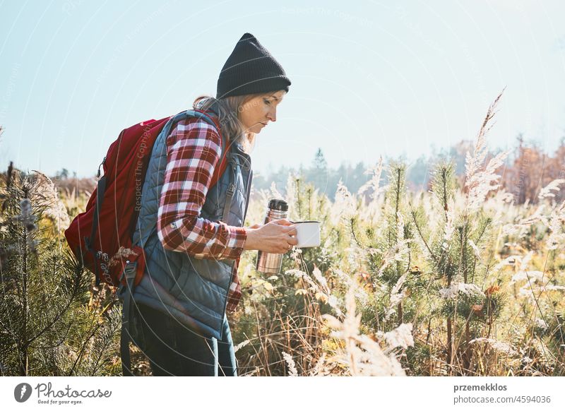 Woman taking a break and relaxing in the bright warm sunlight with a cup of coffee during a summer outing. Woman standing on the path and looking away. Woman with backpack hiking through tall grass along path in mountains