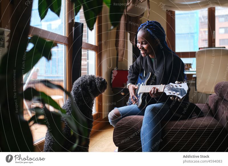 Black woman playing guitar sitting on couch opposite poodle dog pet living room musician sofa companion jeans happy animal friend owner loyal free time